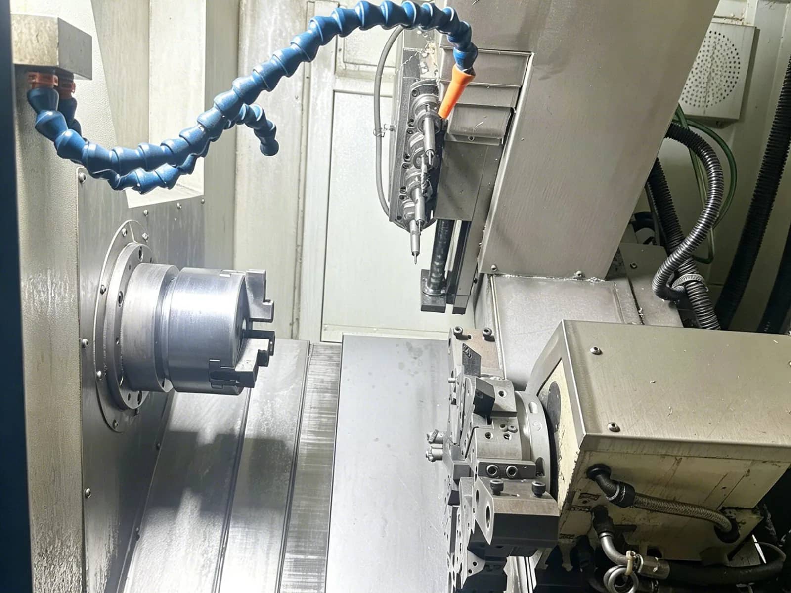 Turn-mill machine cabin showing a three-jaw chuck, live-tool turret, blue coolant hose, and metal chips on the way covers